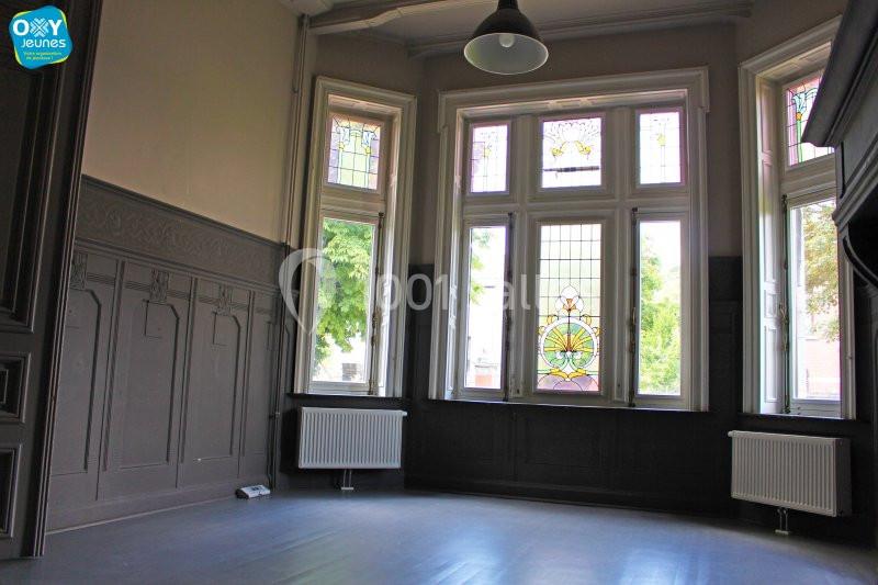 Salle vide avec parquet sombre, murs ornés de moulures et grandes fenêtres avec vitraux colorés laissant entrer la lumière.