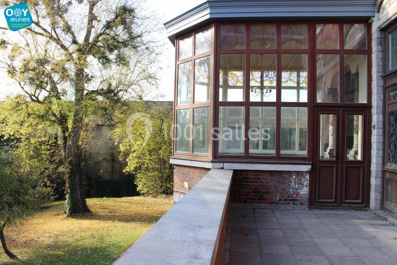 Véranda vitrée donnant sur un jardin arboré avec pelouse, vue depuis une terrasse en pierre.
