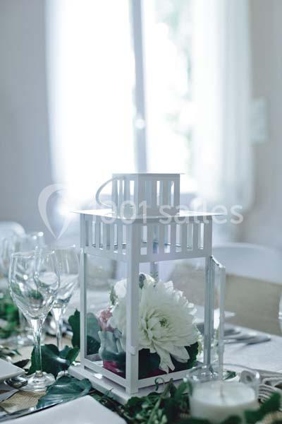 Lanterne blanche décorative contenant des fleurs blanches, posée sur une table dressée avec verres et feuillage.