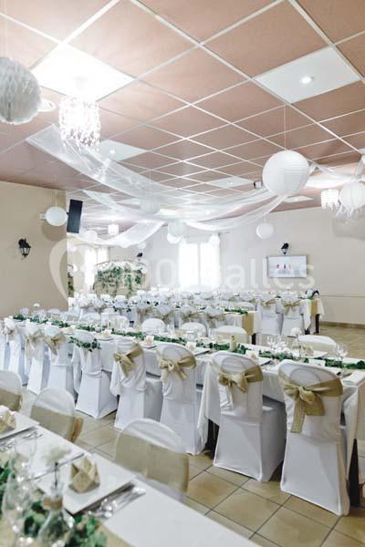 Salle de réception décorée avec des tables alignées, nappes blanches, chaises ornées de nœuds dorés et guirlandes suspendues.