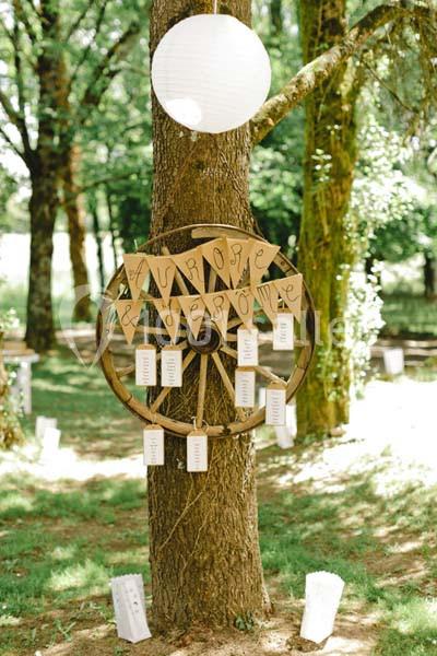 Roue en bois accrochée à un arbre, décorée de cartons suspendus et d'une lanterne blanche dans un cadre boisé.