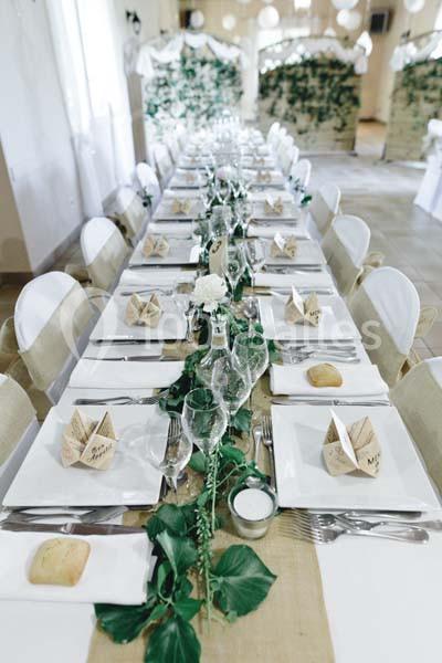 Table de réception décorée avec une nappe en toile de jute, feuillage, vaisselle blanche et pliages en papier.