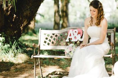 Une mariée en robe blanche est assise sur un banc en bois décoré des lettres ’A&J’, tenant un bouquet de fleurs.
