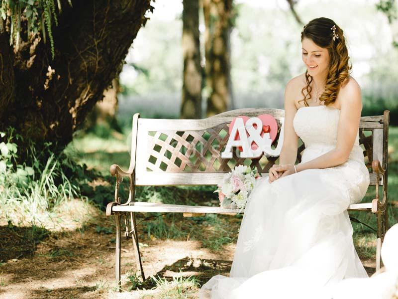 Une mariée en robe blanche est assise sur un banc en bois décoré des lettres ’A&J’, tenant un bouquet de fleurs.