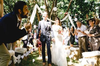 Une mariée en robe blanche est assise sur un banc en bois décoré des lettres ’A&J’, tenant un bouquet de fleurs.