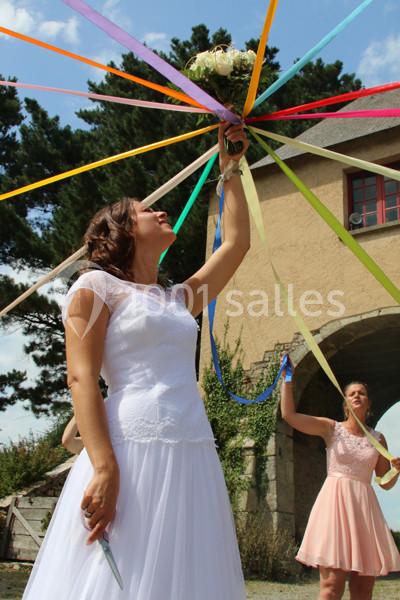 Une mariée tient un bouquet relié à des rubans colorés, entourée d'invités devant un bâtiment en pierre.