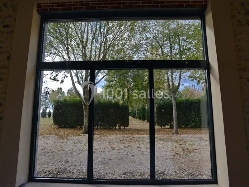 Vue à travers une grande fenêtre sur un jardin arboré avec des haies et des arbres en arrière-plan.
