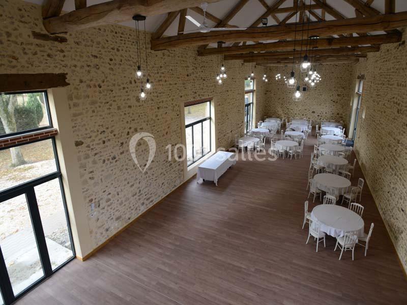 Salle spacieuse avec murs en pierre, poutres apparentes, parquet, grandes fenêtres et tables rondes dressées.