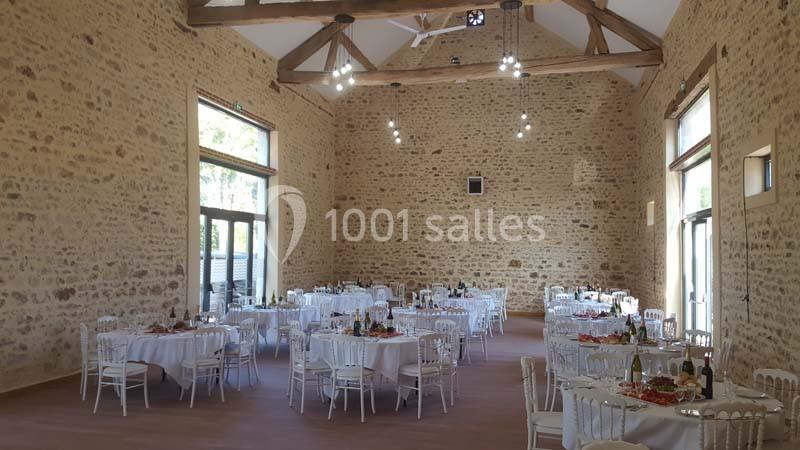 Salle de réception en pierre avec tables rondes dressées pour un repas, éclairée par des suspensions et la lumière naturelle.