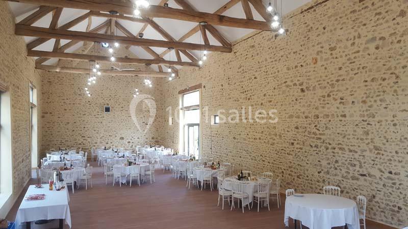 Salle de réception lumineuse avec murs en pierre, poutres apparentes et tables rondes dressées pour un événement.