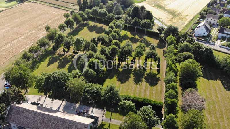 Vue aérienne d'un jardin arboré avec des allées herbeuses, entouré de champs et de quelques bâtiments résidentiels.