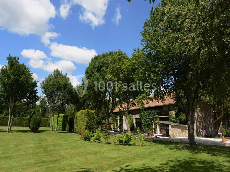 Jardin verdoyant avec pelouse, arbres et une maison en pierre partiellement couverte de végétation sous un ciel bleu.