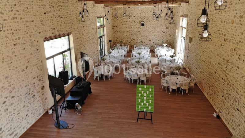Salle de réception en pierre avec tables rondes dressées, grandes fenêtres et plafond avec poutres apparentes.