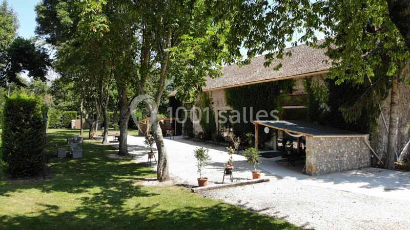 Bâtiment en pierre entouré d'arbres, avec une terrasse ombragée et un chemin gravillonné dans un cadre verdoyant.
