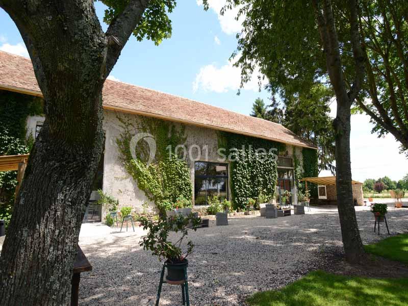 Bâtiment en pierre couvert de lierre, entouré d'arbres et d'une cour gravillonnée sous un ciel ensoleillé.