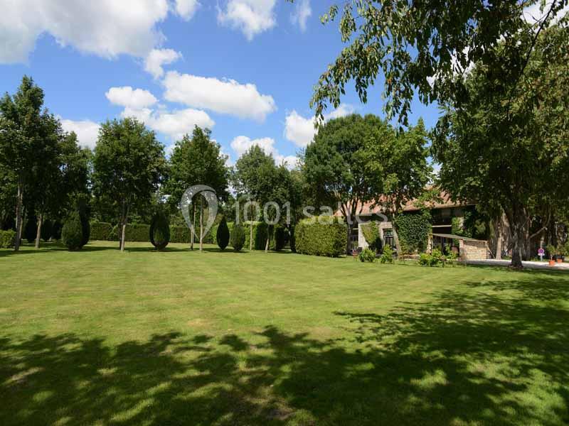 Jardin verdoyant avec pelouse, arbres alignés et une maison en pierre partiellement visible sous un ciel bleu.