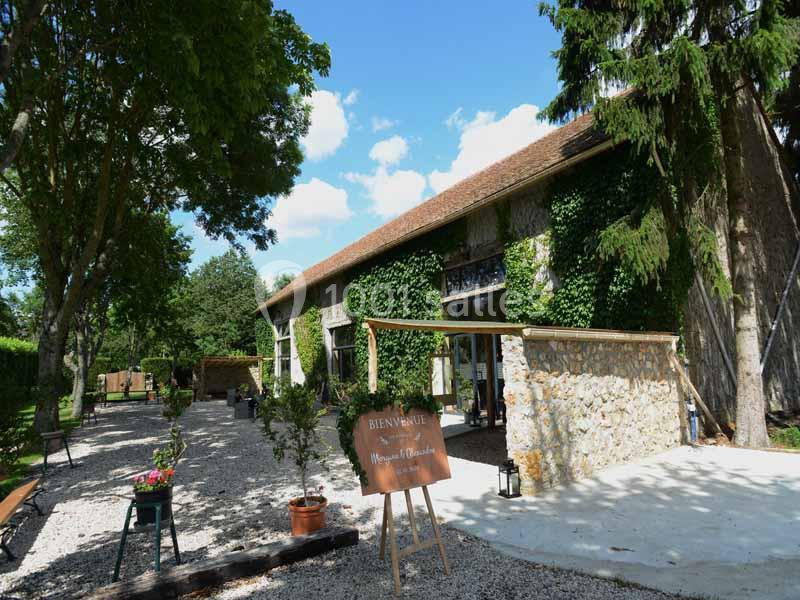 Façade d'une bâtisse en pierre couverte de végétation, entourée d'arbres et d'une cour gravillonnée avec un panneau d…