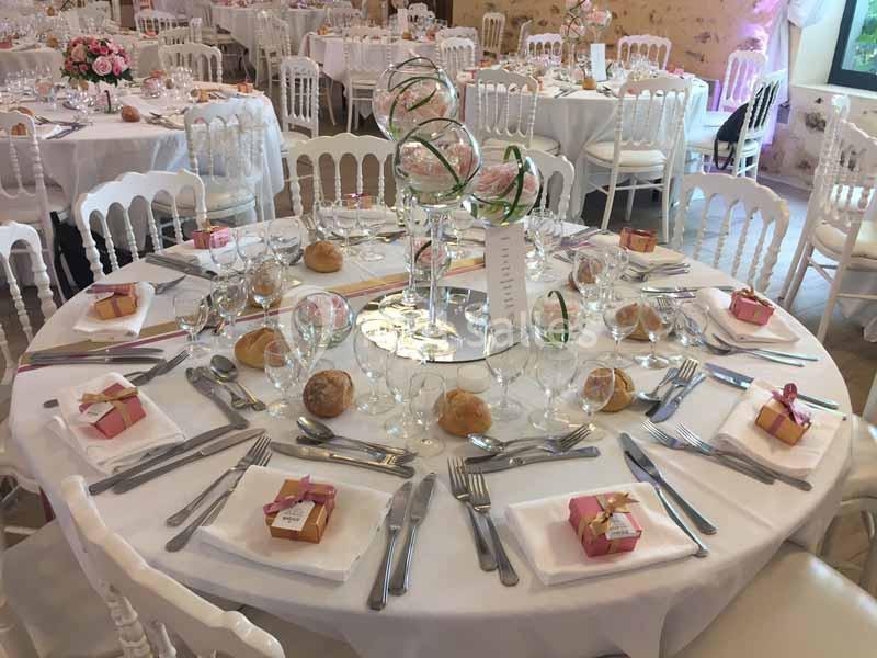 Table de réception élégamment dressée avec nappes blanches, couverts, verres, petits cadeaux et décoration florale centrale.