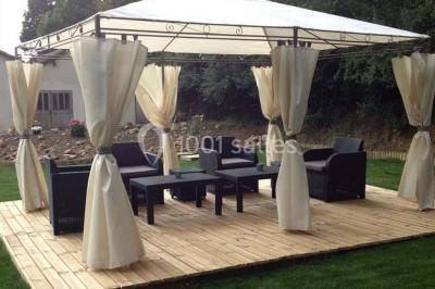 Salle lumineuse avec grandes fenêtres, mobilier en rotin gris et poutres apparentes, donnant sur un espace extérieur…