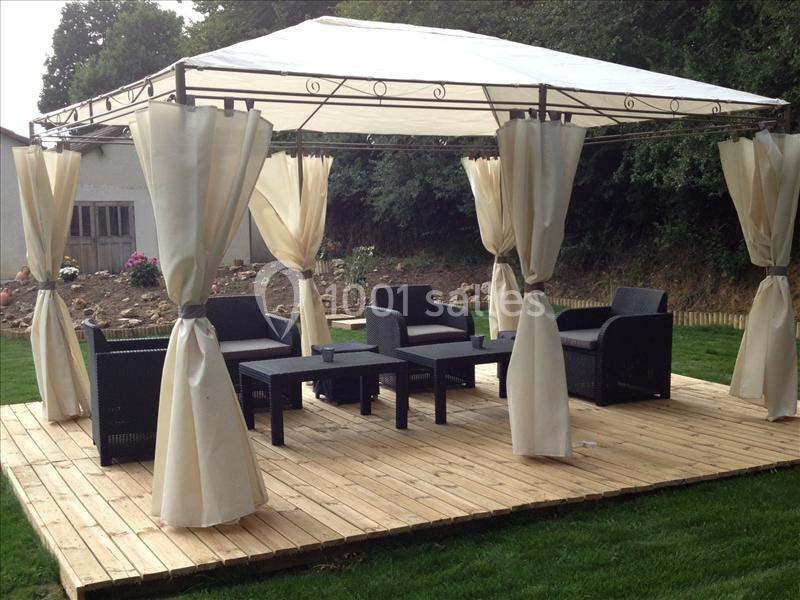 Tonnelle avec rideaux ouverts, mobilier de jardin en rotin noir et table basse sur une terrasse en bois dans un jardin.