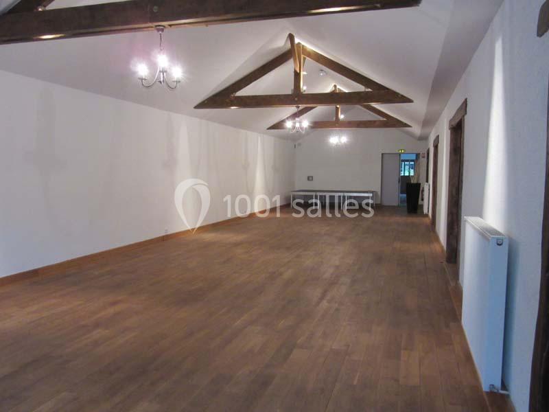 Salle vide avec parquet en bois, murs blancs, poutres apparentes au plafond et tables alignées au fond.