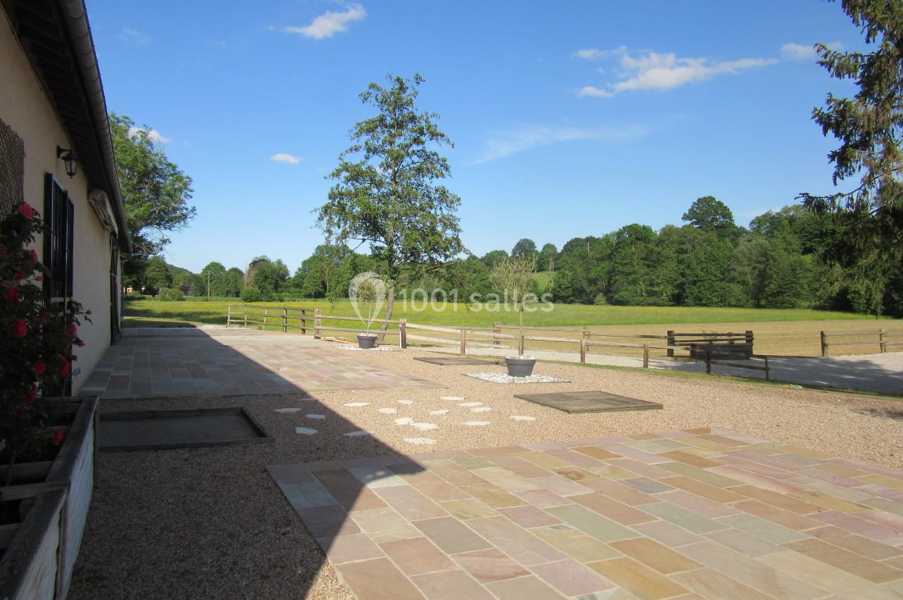 Cour en pavés colorés bordée d'une maison et d'un jardin, avec vue sur un champ et des arbres sous un ciel bleu.