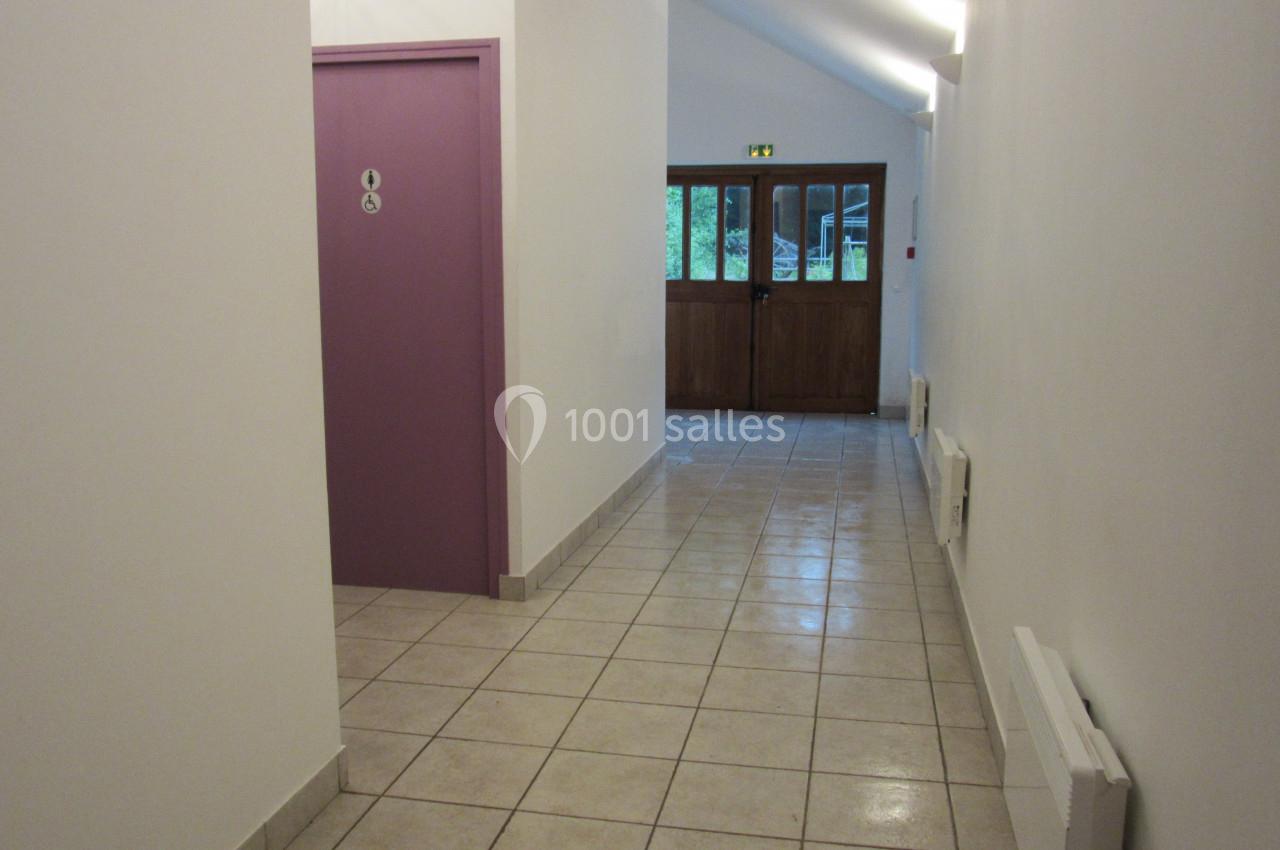 Couloir lumineux avec sol carrelé, porte violette à gauche indiquant des toilettes, et porte en bois au fond.