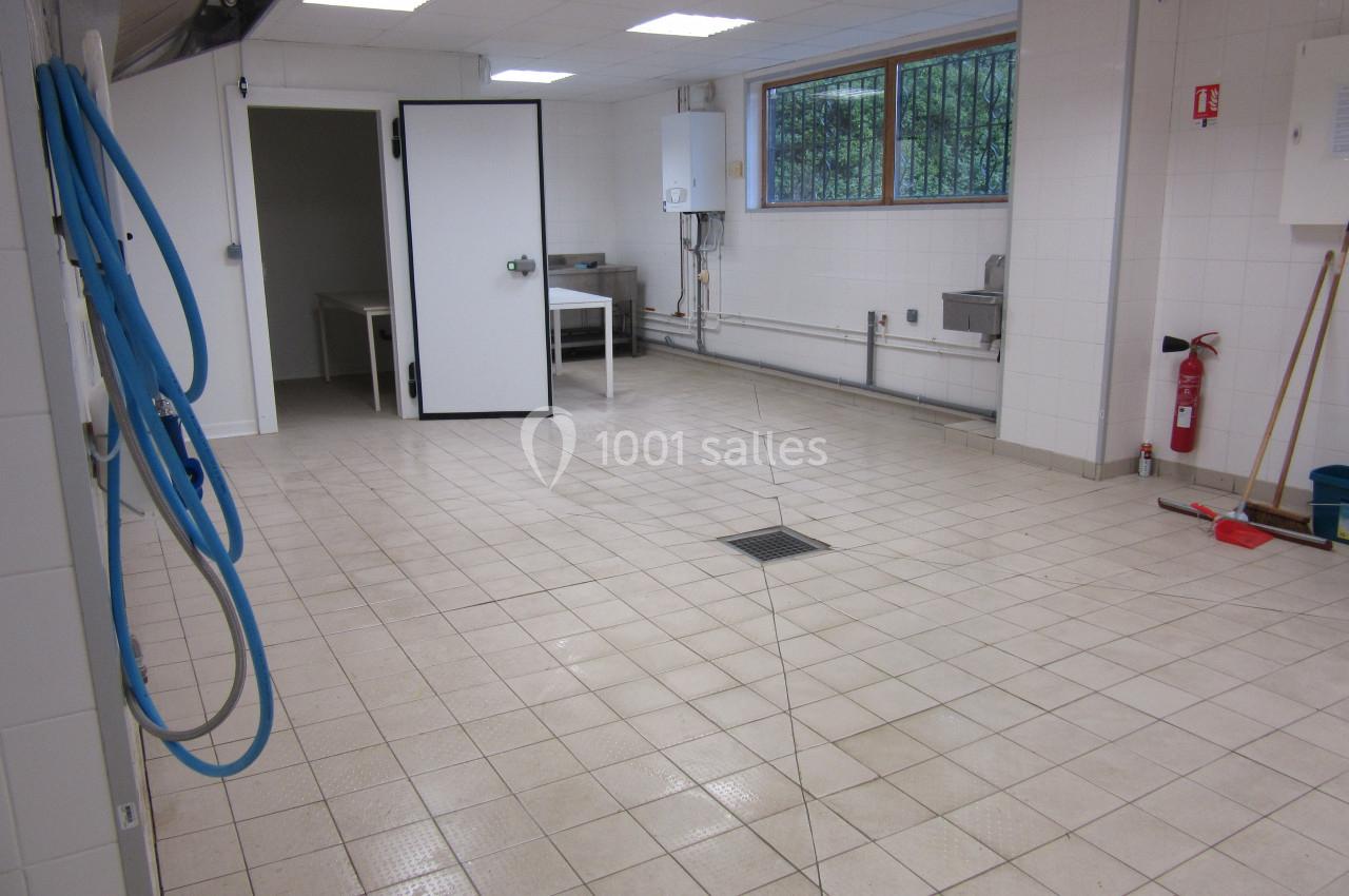 Salle carrelée vide avec murs blancs, éclairage au plafond, porte ouverte, tuyaux apparents et équipements de nettoyage.