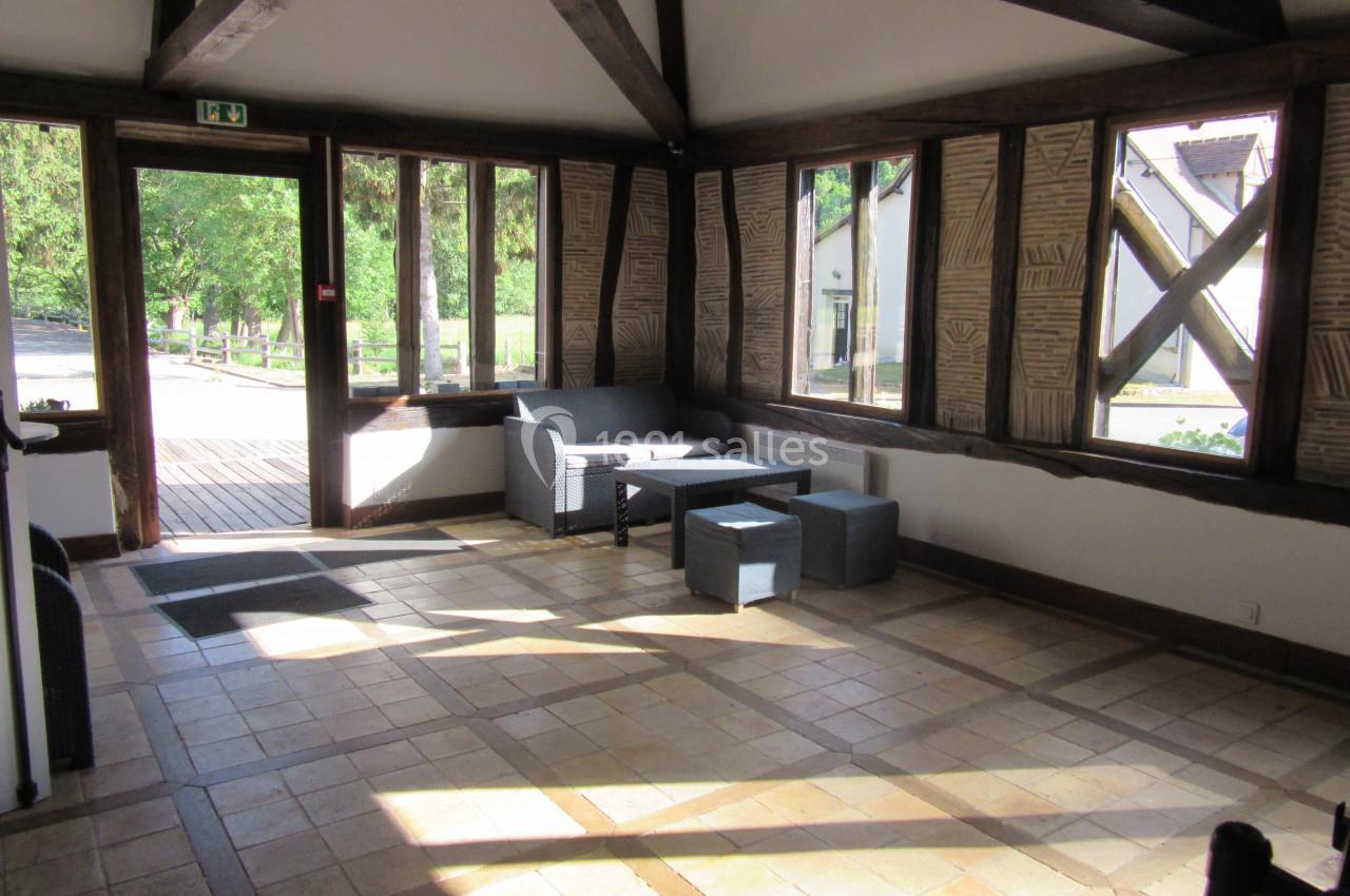 Salle lumineuse avec grandes fenêtres, mobilier en rotin gris et poutres apparentes, donnant sur un espace extérieur…