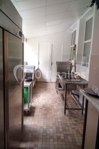 Cuisine professionnelle avec évier inox, réfrigérateur, carrelage au sol et étagères de rangement.