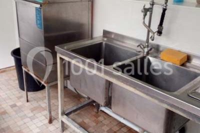 Évier double en inox avec robinet flexible, lave-vaisselle professionnel et sol en carrelage beige dans une cuisine.