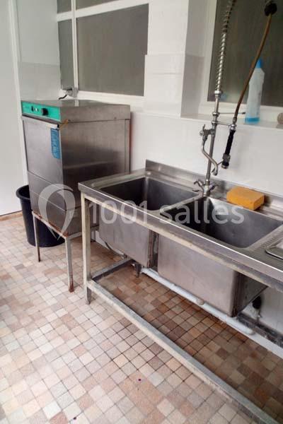 Évier double en inox avec robinet flexible, lave-vaisselle professionnel et sol en carrelage beige dans une cuisine.