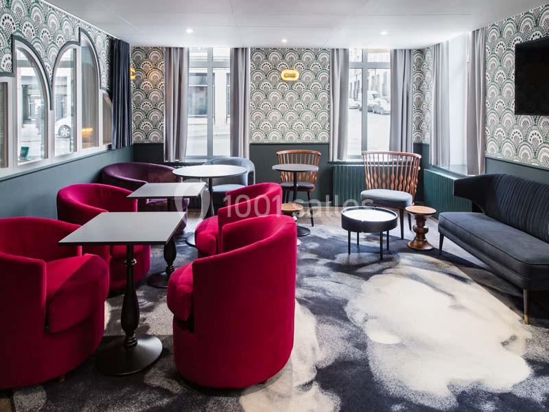Salle de détente avec fauteuils rouges, tables basses, canapé bleu et fenêtres laissant entrer la lumière naturelle.