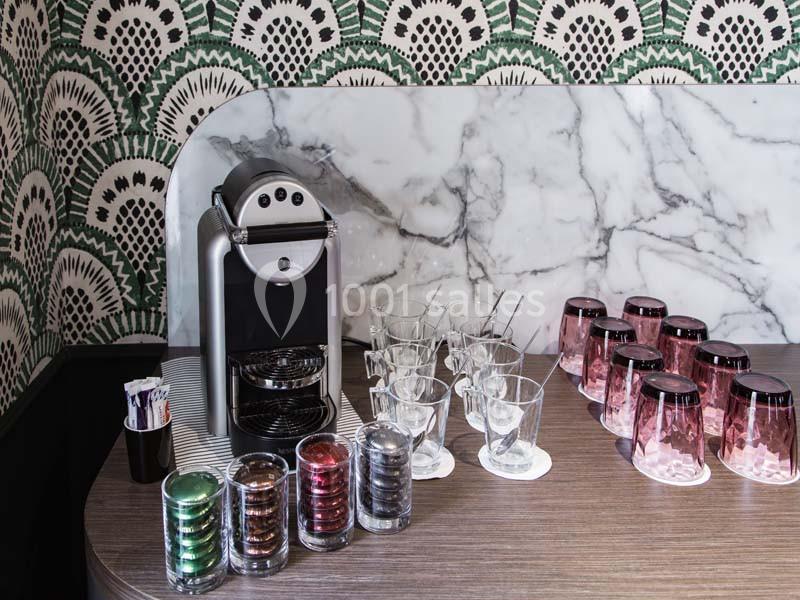 Machine à café avec capsules colorées, verres transparents et gobelets roses sur un comptoir en bois.