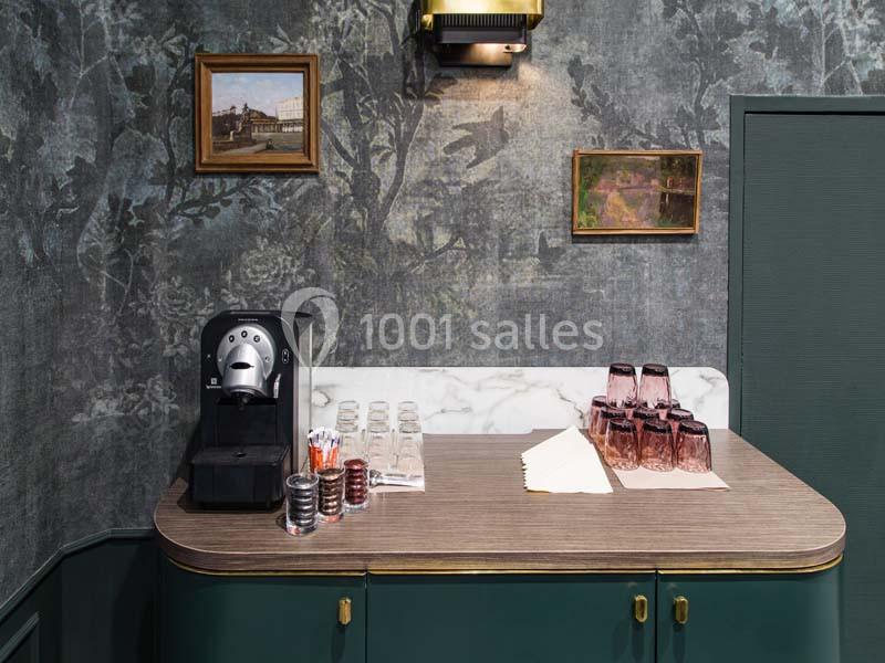 Machine à café avec capsules, verres et serviettes sur un comptoir en bois, décoré de tableaux et d'un mur texturé.