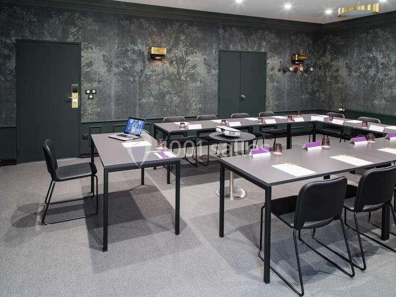 Salle de réunion équipée de tables en U, chaises noires, projecteur et ordinateur portable sur une table.