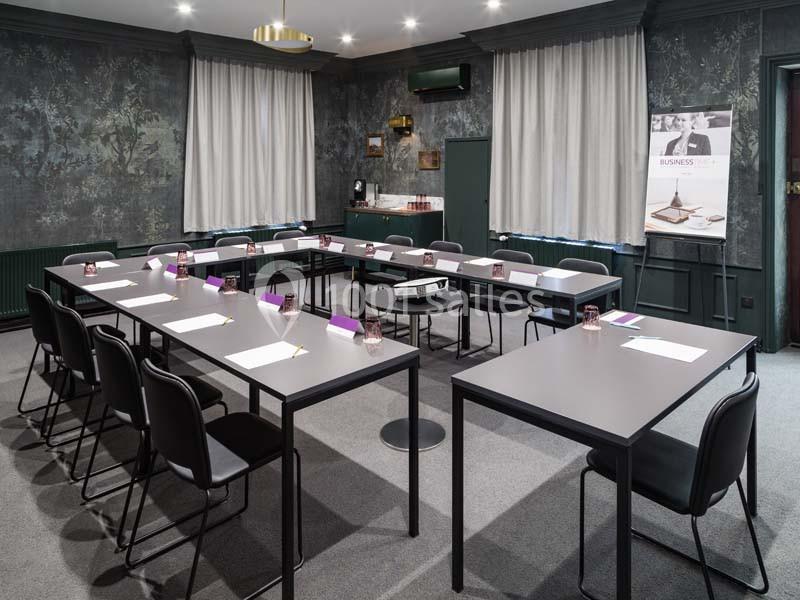 Salle de réunion avec tables disposées en U, chaises noires, rideaux clairs et mur décoré de motifs sombres.