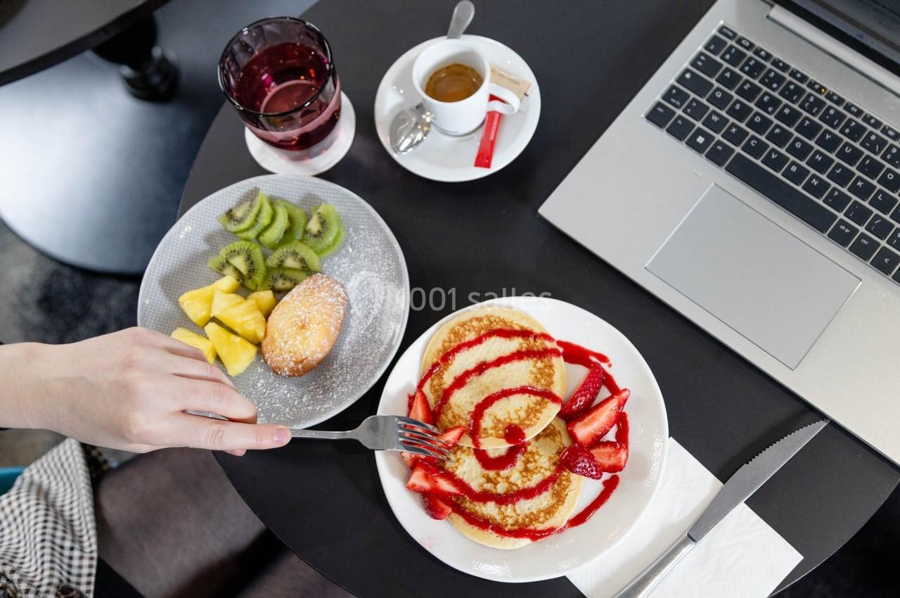 Assiette de pancakes garnis de fraises et coulis, accompagnée de fruits, café et ordinateur sur une table noire.