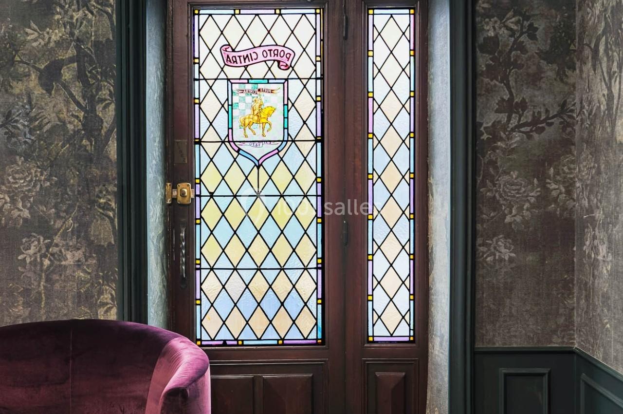 Porte en bois avec vitraux colorés, encadrée par un mur sombre et un fauteuil violet au premier plan.