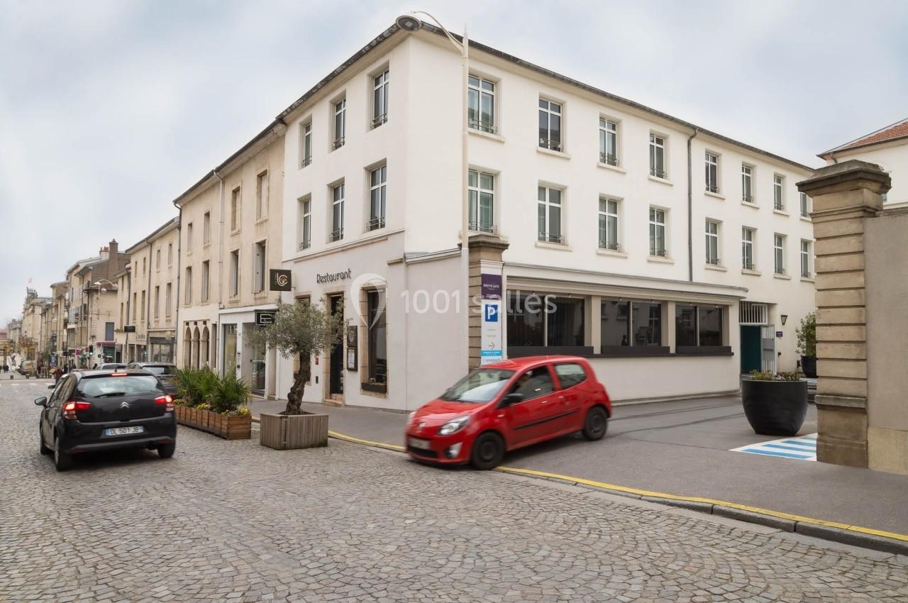 Façade d'un bâtiment moderne avec des voitures stationnées et circulant sur une rue pavée.