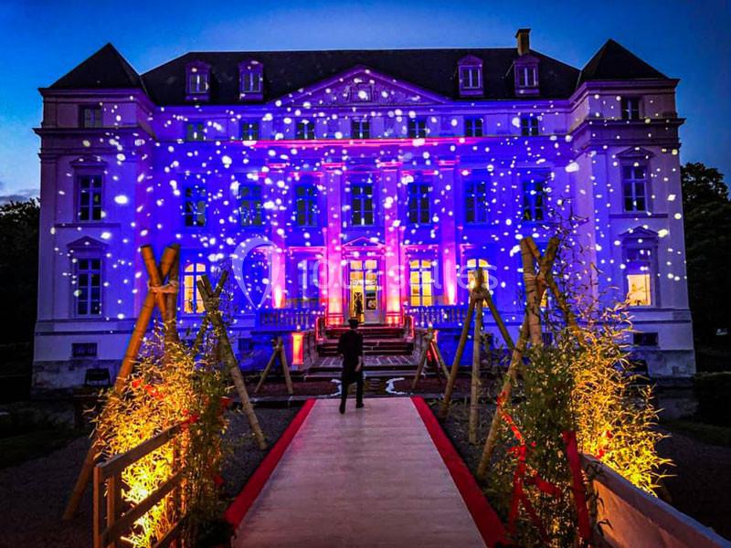 Façade d'un château illuminé par des projections colorées, vue depuis une passerelle bordée de lumières.