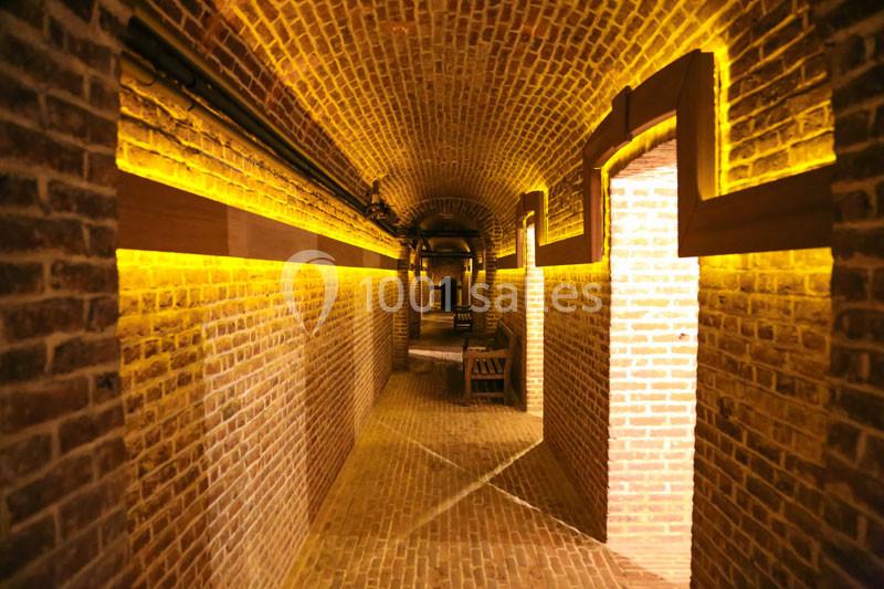 Couloir voûté en briques éclairé par des lumières jaunes, avec des bancs en bois disposés le long des murs.
