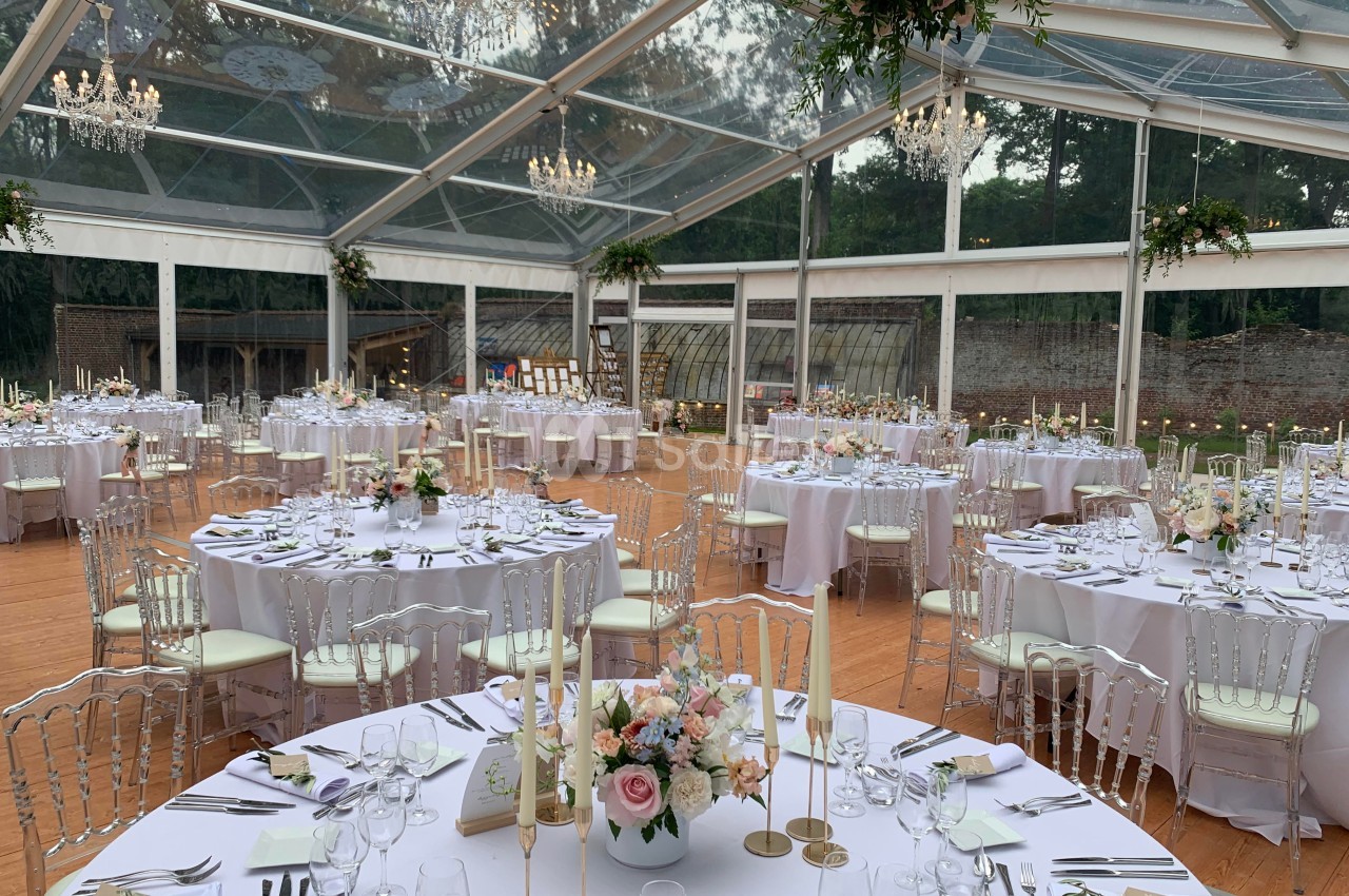 Salle de réception sous une tente transparente, tables rondes décorées de fleurs et chandeliers suspendus.
