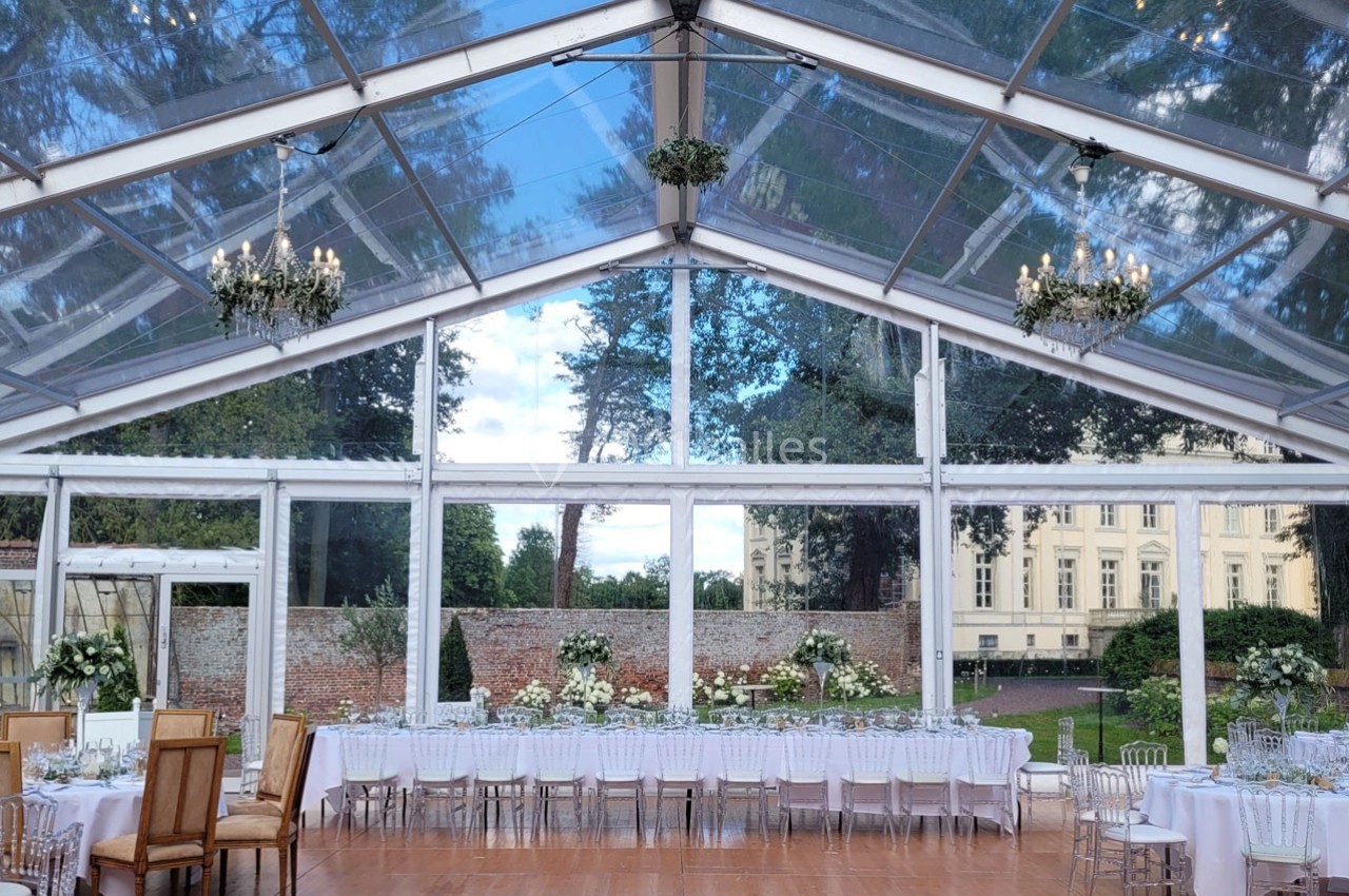 Salle de réception sous une tente transparente avec tables décorées, chaises élégantes et vue sur un jardin et un bâtiment…