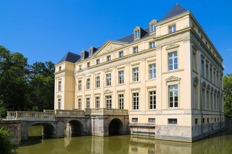 Façade d'un château classique beige avec pont et douves, entouré de végétation sous un ciel bleu.