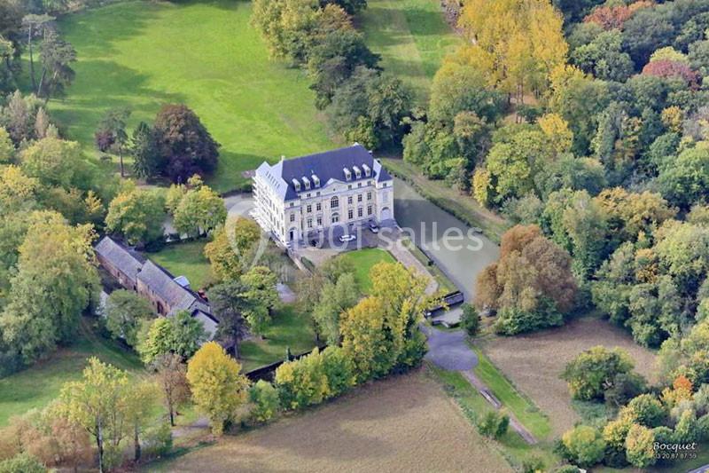 Vue aérienne d'un château entouré de verdure, avec un plan d'eau adjacent et des bâtiments annexes à proximité.