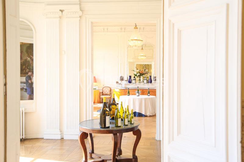 Table ronde avec des bouteilles de vin dans une pièce lumineuse ornée de moulures et de lustres.