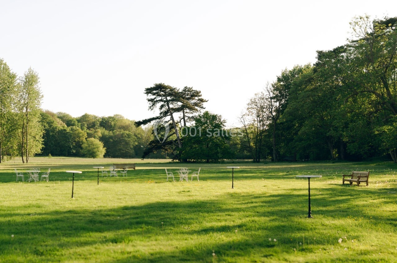 Pelouse verdoyante parsemée de tables et chaises blanches, entourée d'arbres sous un ciel dégagé.