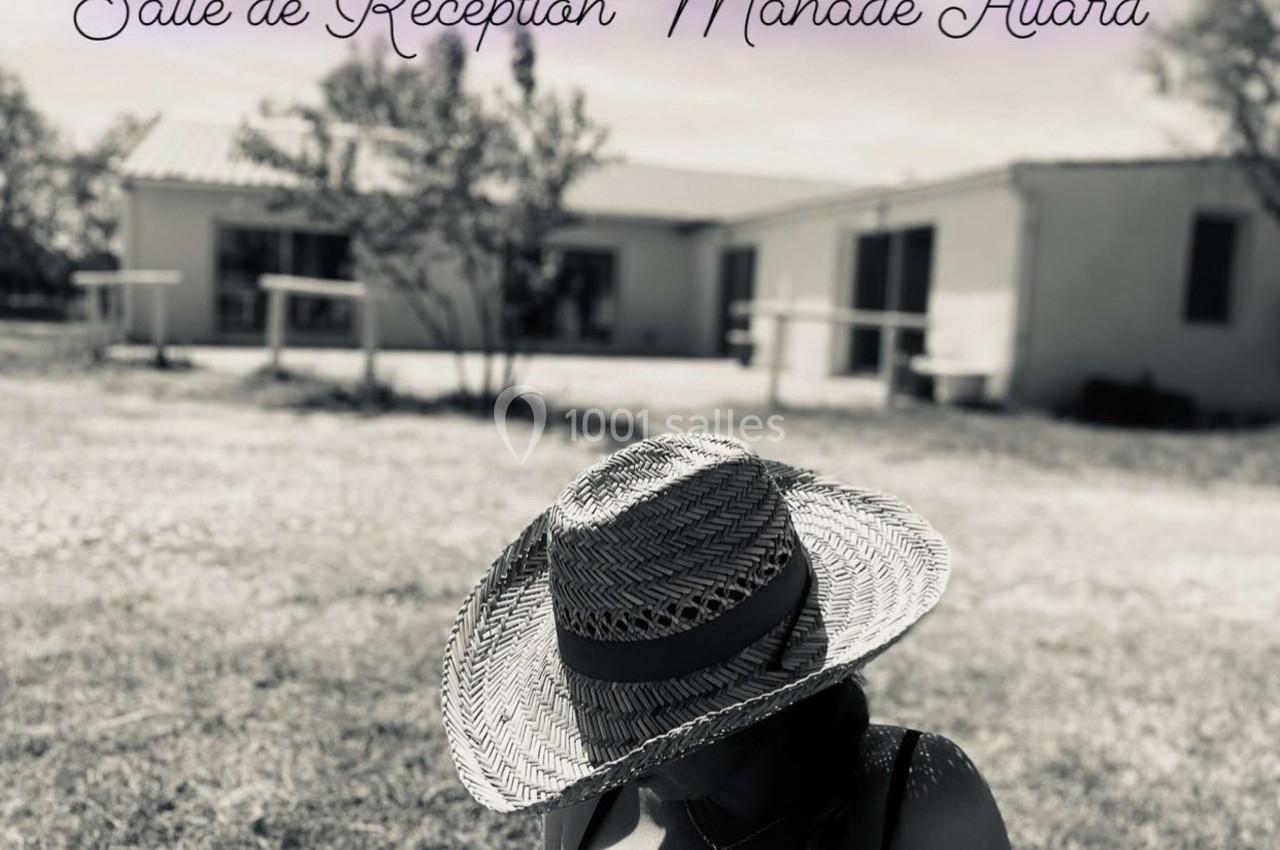 Femme portant un chapeau en paille au premier plan, bâtiment blanc et arbres flous en arrière-plan.