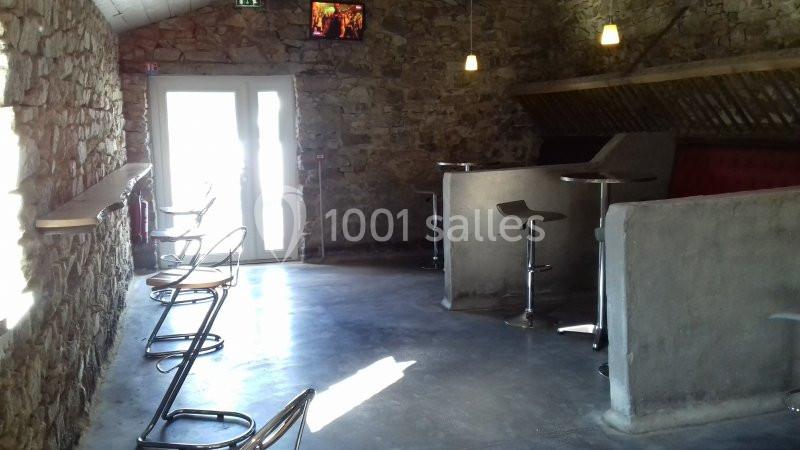 Salle avec murs en pierre, tables hautes, tabourets métalliques et lumière naturelle entrant par une porte vitrée.